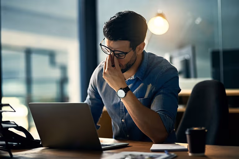 Man in glasses rubbing eyes while sitting at a desk with a laptop, appearing fatigued in an office setting.