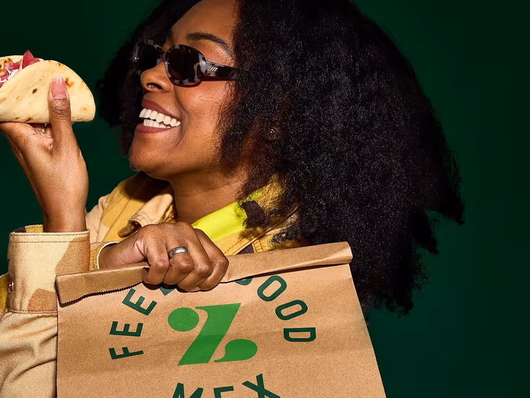 Woman in sunglasses smiling while holding a Zambrero Fat Taco in one hand and a brown paper Zambrero bag in the other