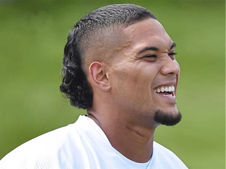 Man with a skullet haircut featuring a fade and curly mullet, smiling outdoors in bright lighting.