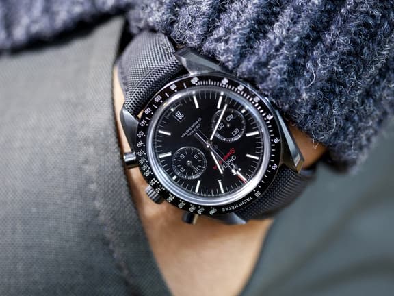 Black Omega Speedmaster watch on wrist with textured strap and dark sweater.