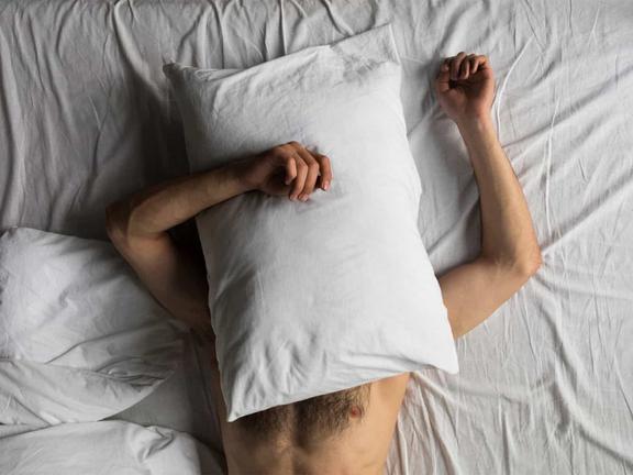 Naked man lying on a bed with a pillow on his face