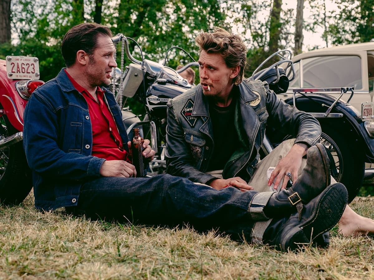 Austin Butler and Tom Hardy sit on grass with motorcycles in the background, wearing leather jackets and casual attire.