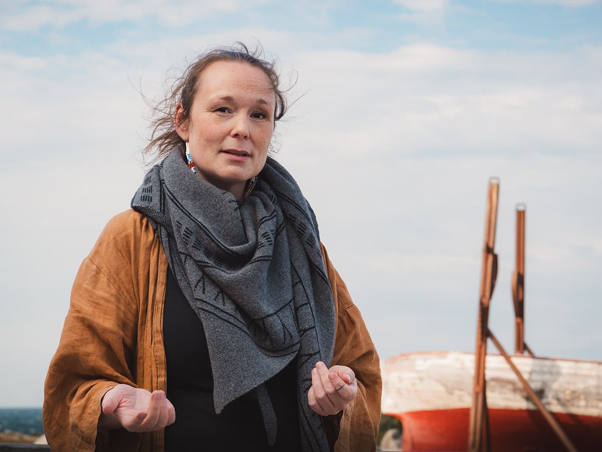 Ujammiugaq Engell stands outdoors wearing a grey scarf and brown coat, with a boat in the background.
