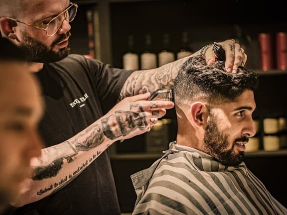 Barber with tattoos trims a man's hair in a Sydney barbershop, with shelves of grooming products in the background.