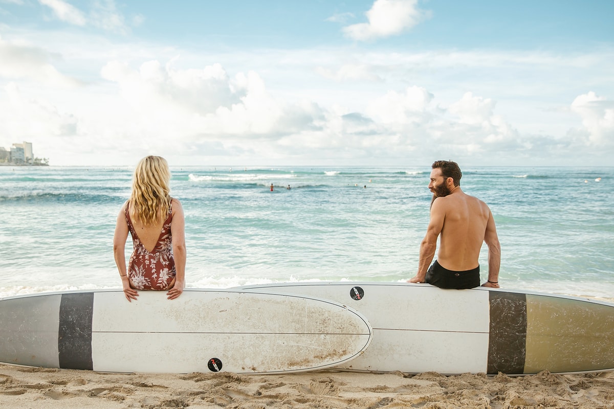 Backs of Jimmy and Emma sitting on edges of their surfboard standing on sides in beach sand