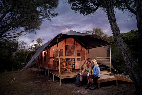 Man and woman sitting on a wooden platform in front of cabin-like elevated tent