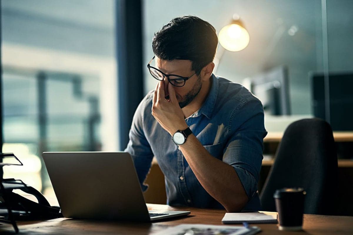 Man in glasses rubbing eyes while sitting at a desk with a laptop, appearing fatigued in an office setting.