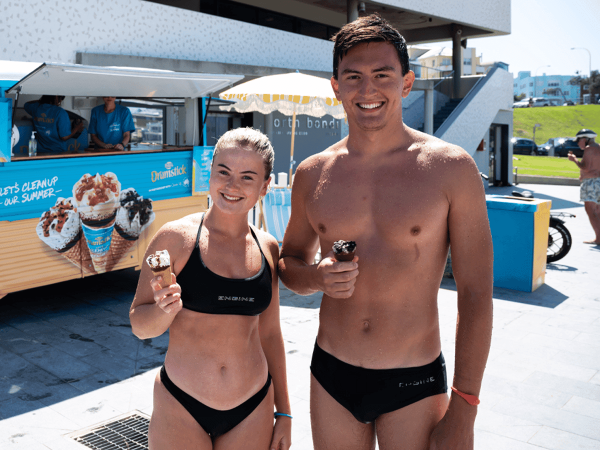 Man and woman in swimwear holding a drumstick with ice-cream truck in the background