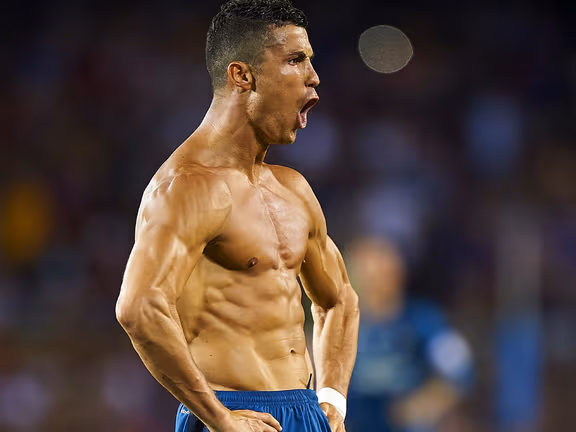 Cristiano Ronaldo shirtless, flexing muscles on a football field with intense expression.