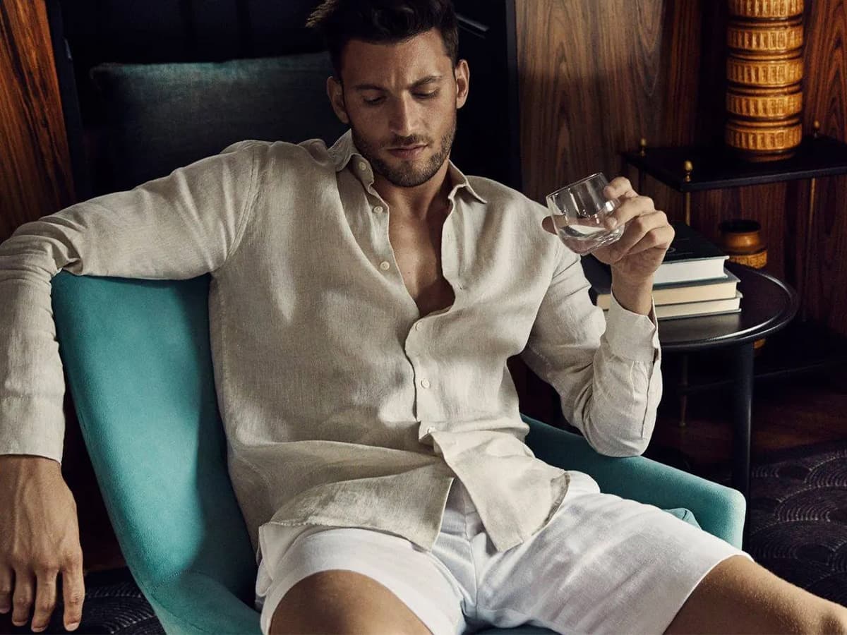 Man in off-white shirt and white shorts sitting on a chair holding a drink