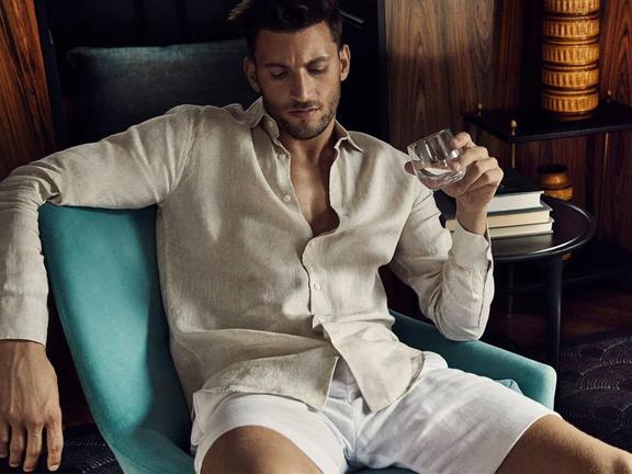 Man in off-white shirt and white shorts sitting on a chair holding a drink
