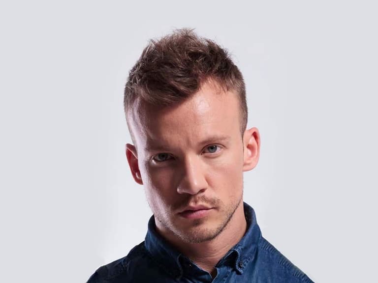 Man with faux hawk hairstyle and receding hairline wearing a blue denim shirt against a grey background.