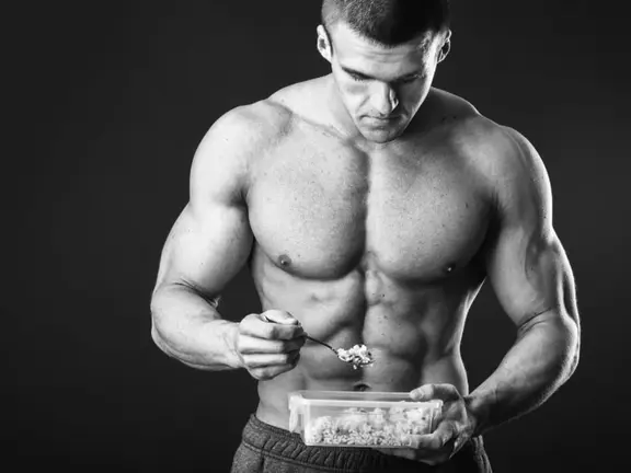 Greyscale image of a shirtless muscular man eating from a lunchbox he’s holding in one hand