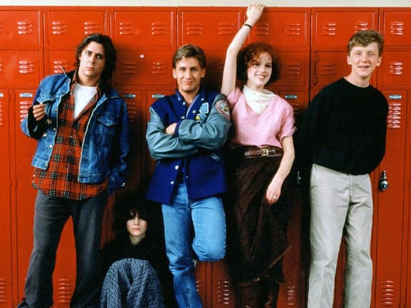 Five teenagers in '80s fashion stand against red lockers; denim, plaid, and varsity jacket styles visible.