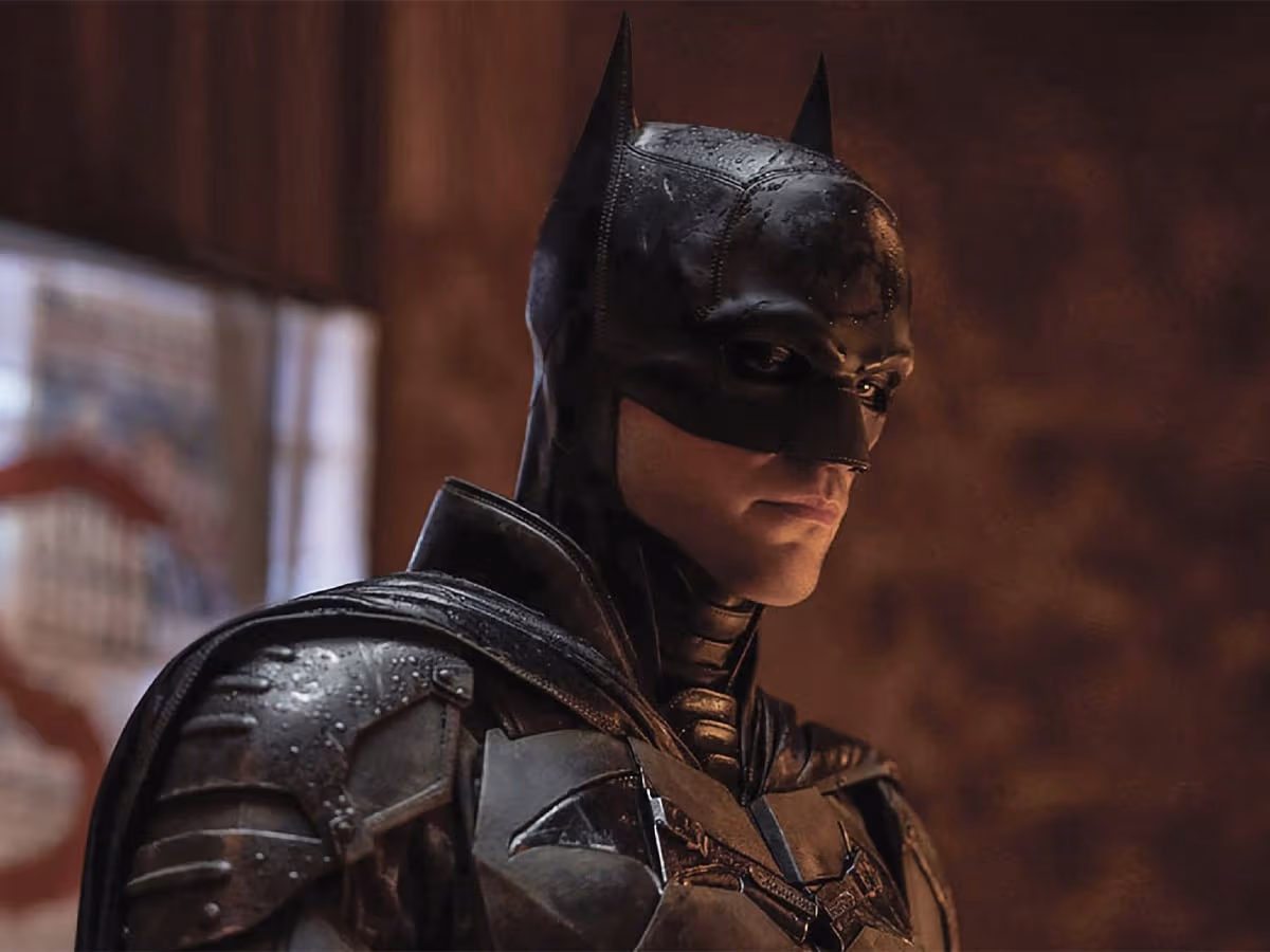 Batman in dark suit stands in dimly lit room, showcasing detailed armour and iconic cowl.