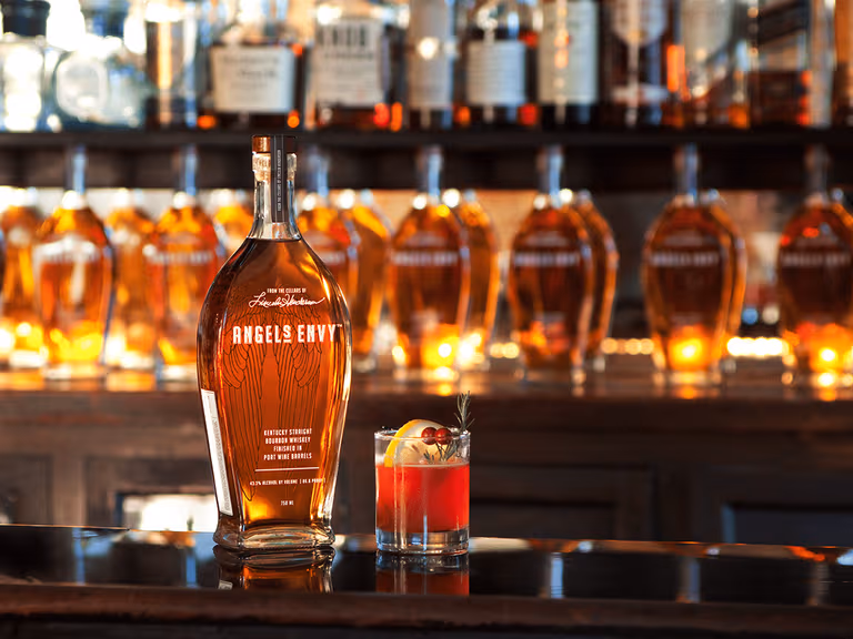 Angel's Envy bottle and cocktail on a bar with backlit whisky bottles.