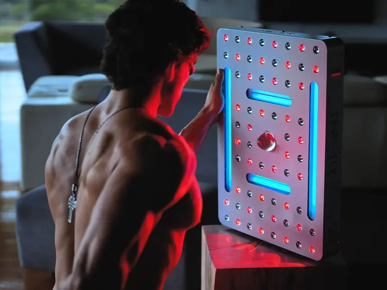 Muscular man using a red and blue light therapy panel indoors, facing the device with his back exposed, illuminated by red light