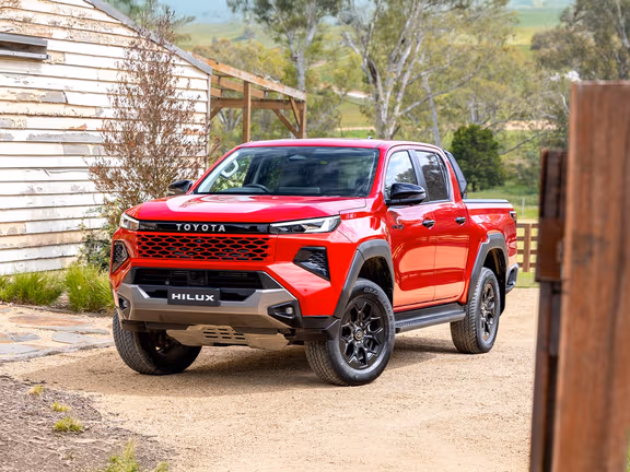 2026 Toyota HiLux Rogue in red, front three-quarter view, parked on a rural dirt driveway with a wooden building nearby.