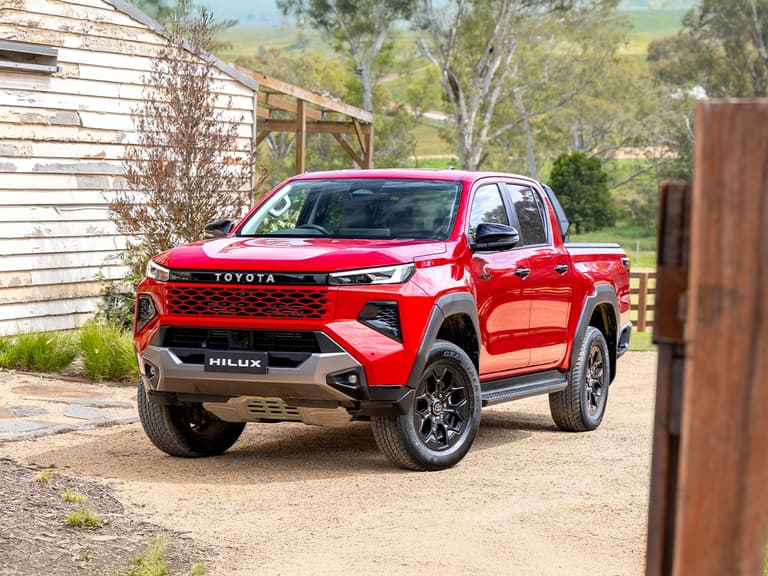 2026 Toyota HiLux Rogue in red, front three-quarter view, parked on a rural dirt driveway with a wooden building nearby.