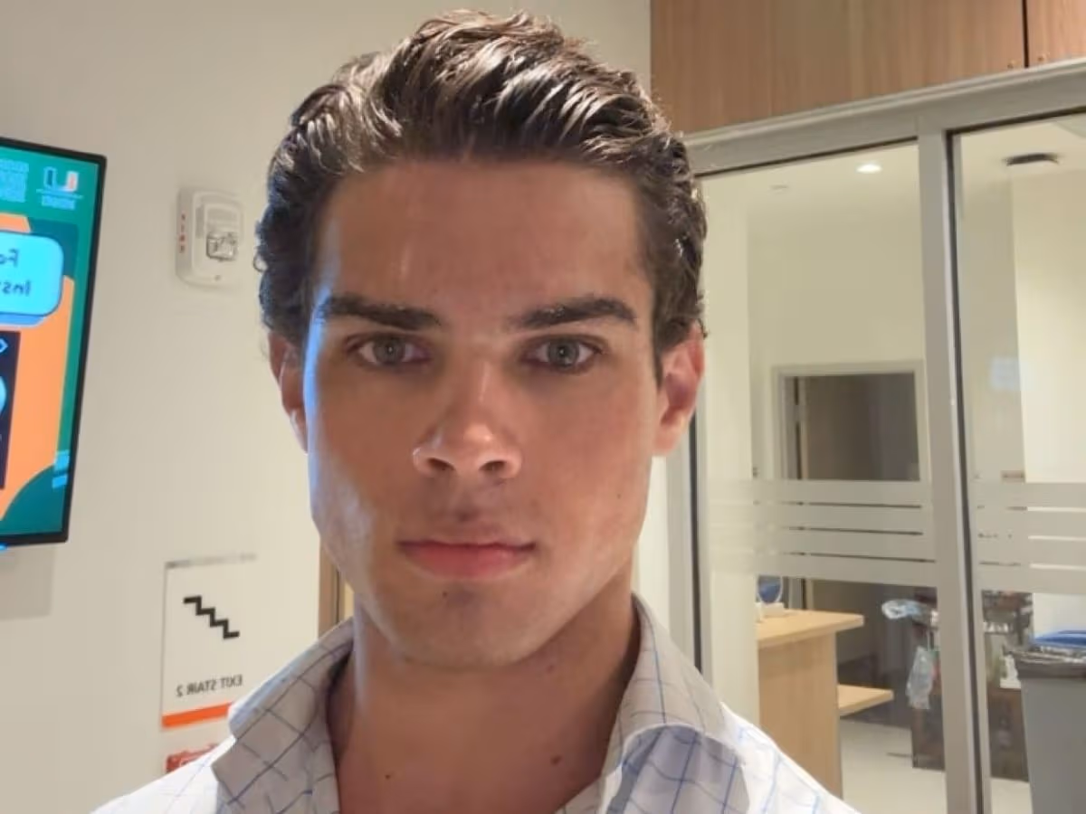 Man with slicked-back hair and a serious expression stands indoors, wearing a checkered shirt, with office decor in the background.