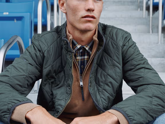 Man wearing a quilted olive Superdry jacket over a hoodie, seated in stadium stands