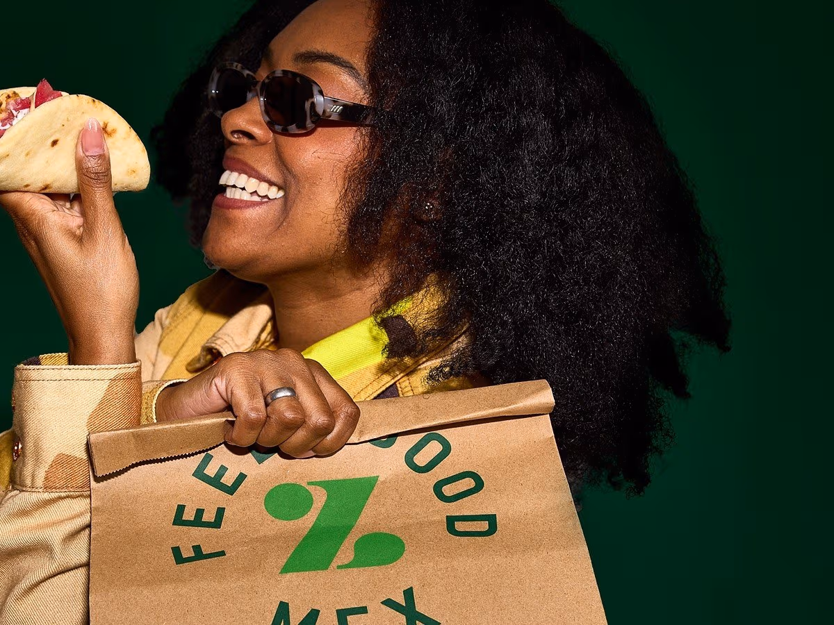 Woman in sunglasses smiling while holding a Zambrero Fat Taco in one hand and a brown paper Zambrero bag in the other