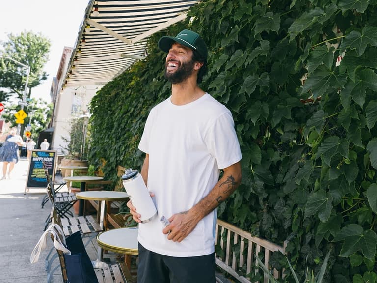 aniel Ricciardo smiling with his eyes closed, wearing a plain white t-shirt and green trucker cap while holding a white water bottle outside a cafe.