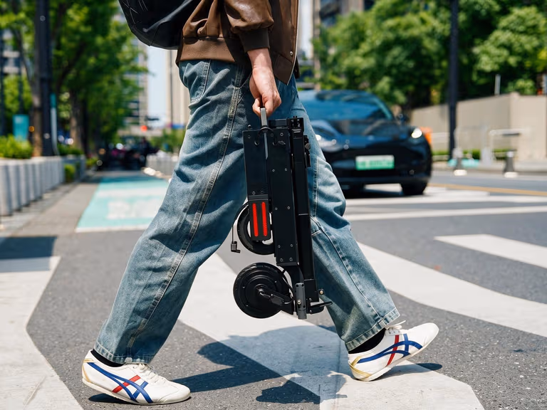 Person carrying the folded Blizwheel F0.5 electric scooter by its handle across a pedestrian crossing