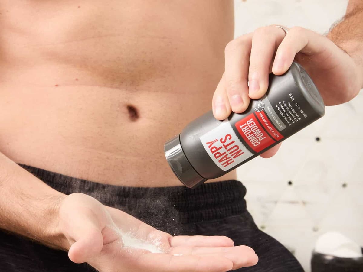 A man tipping Happy Nuts Comfort Powder from its bottle into his open palm, standing in a bathroom in dark shorts.