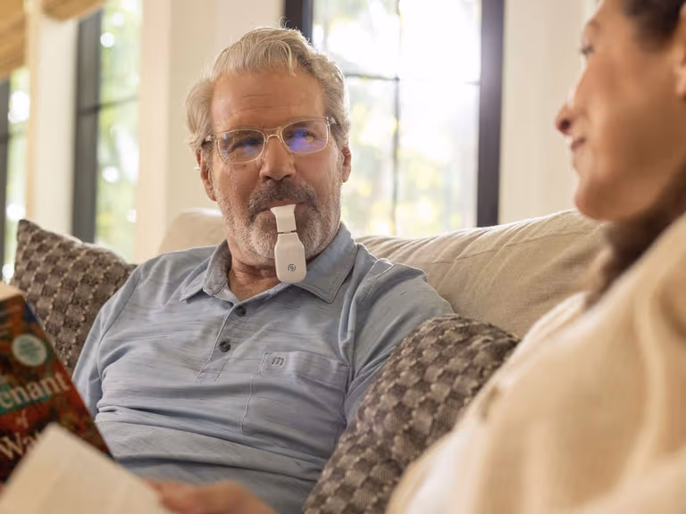 A man using the Spring Sleep eXciteOSA daytime therapy device while reading on the couch beside his partner.