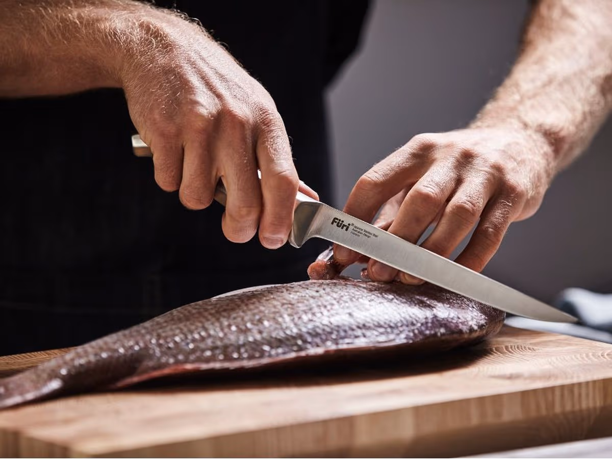 A whole fish being filleted with a Furi filleting knife on a timber chopping board, the blade following the backbone behind the gill plate