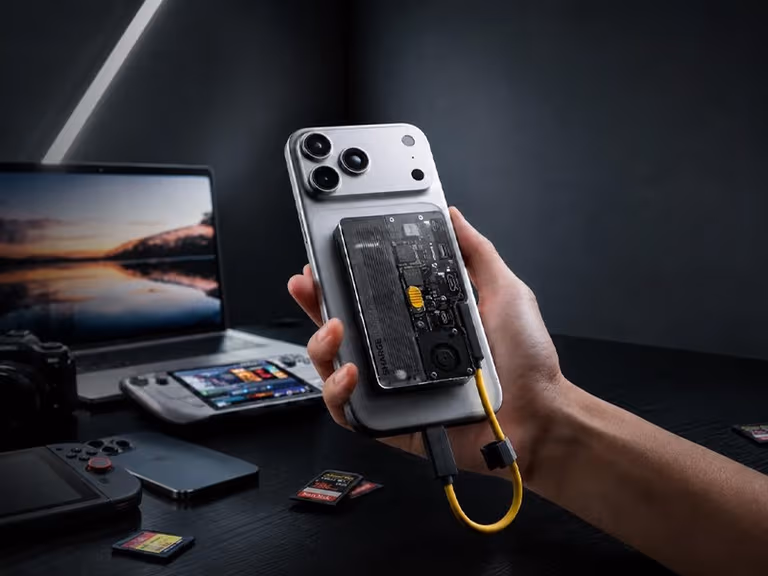 SHARGE Disk Pro 2 magnetically attached to the back of a phone, held above a desk with a laptop, handheld console and memory cards