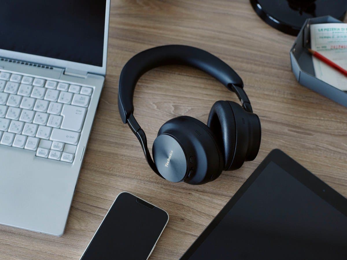 Technics EAH-A1000 wireless headphones in Black on a desk beside a laptop, phone and tablet