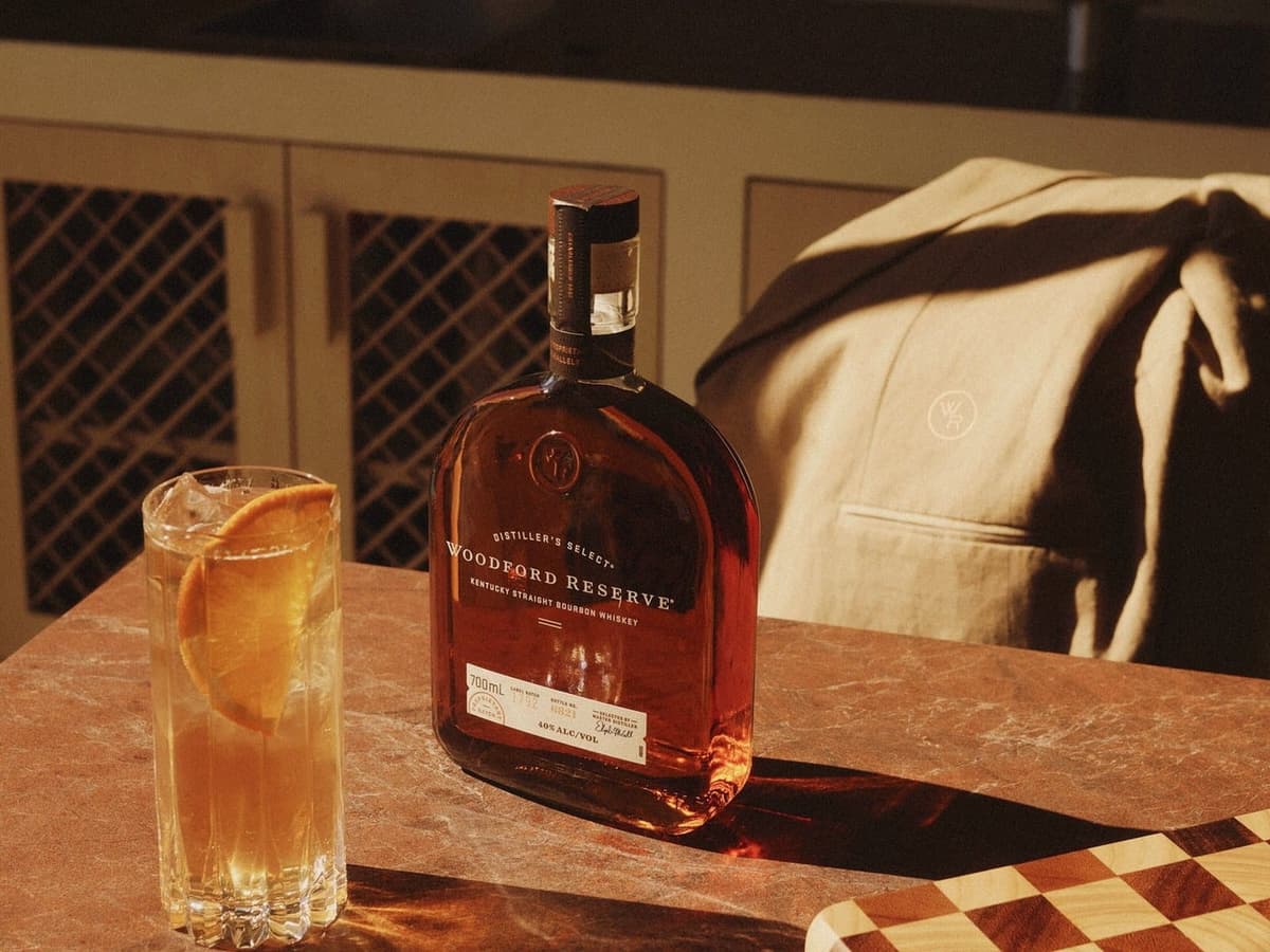 A bottle of Woodford Reserve bourbon and a highball with an orange slice on a stone bench, with the dune linen Host Tux draped over a chair behind.
