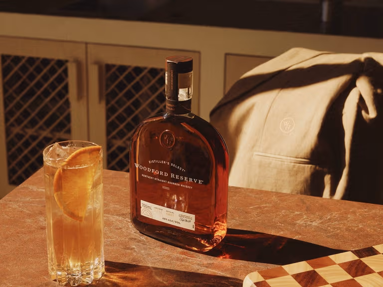 A bottle of Woodford Reserve bourbon and a highball with an orange slice on a stone bench, with the dune linen Host Tux draped over a chair behind.