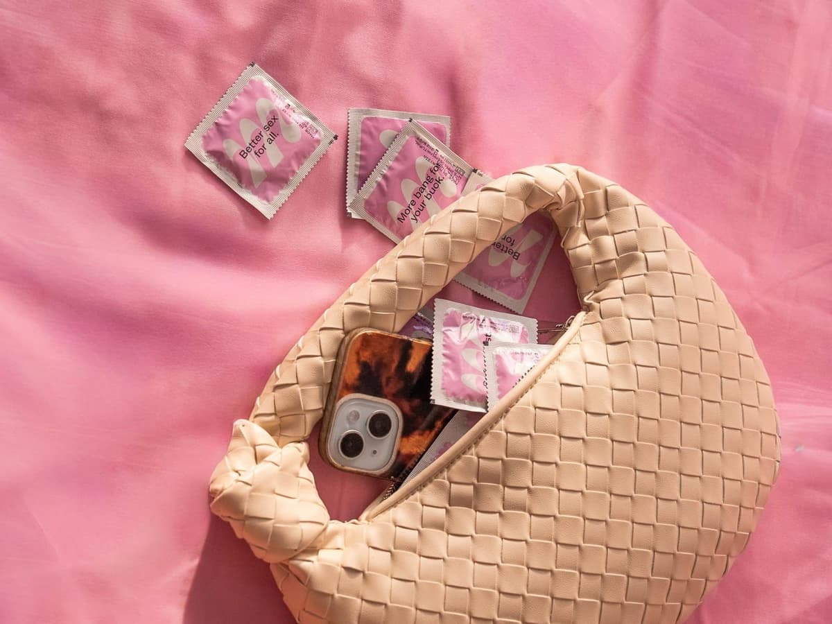 A woven cream handbag on pink sheets with Moments condom sachets and a phone spilling out