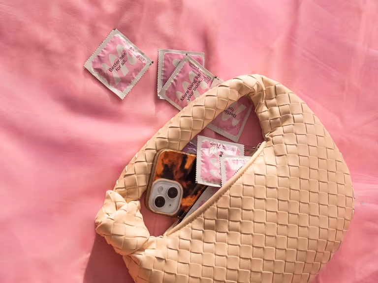 A woven cream handbag on pink sheets with Moments condom sachets and a phone spilling out