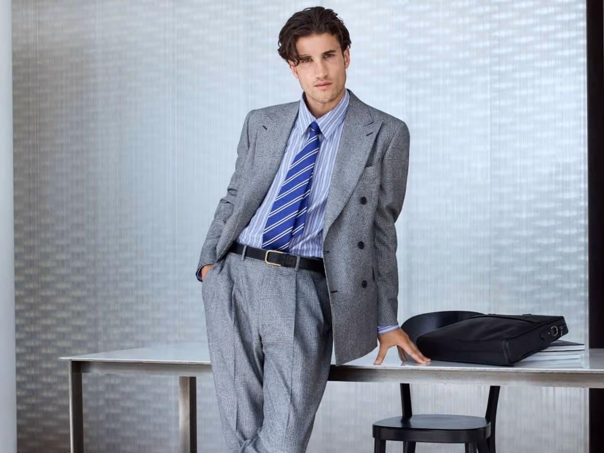 A model in a grey Glen check double-breasted suit, blue striped shirt and striped tie, leaning on a desk beside a black leather briefcase.