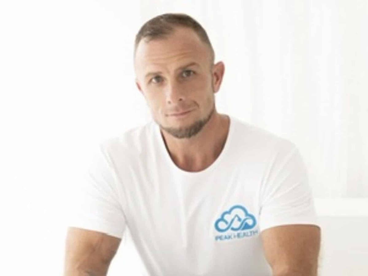 PEAK HEALTH founder Tommy Alsop, in a white PEAK HEALTH T-shirt, seated against a white backdrop with his hands clasped