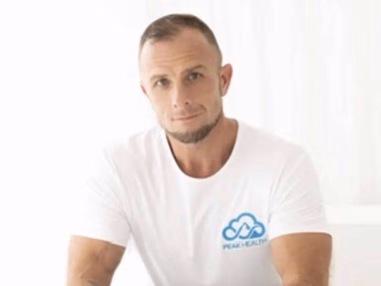 PEAK HEALTH founder Tommy Alsop, in a white PEAK HEALTH T-shirt, seated against a white backdrop with his hands clasped