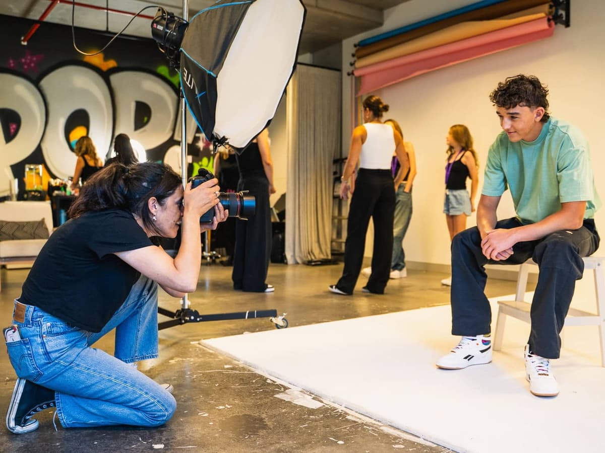 A POP Photography photographer crouches with a camera to shoot a teenage boy seated on a stool against a white studio backdrop, with a large softbox and a graffiti POP mural behind her