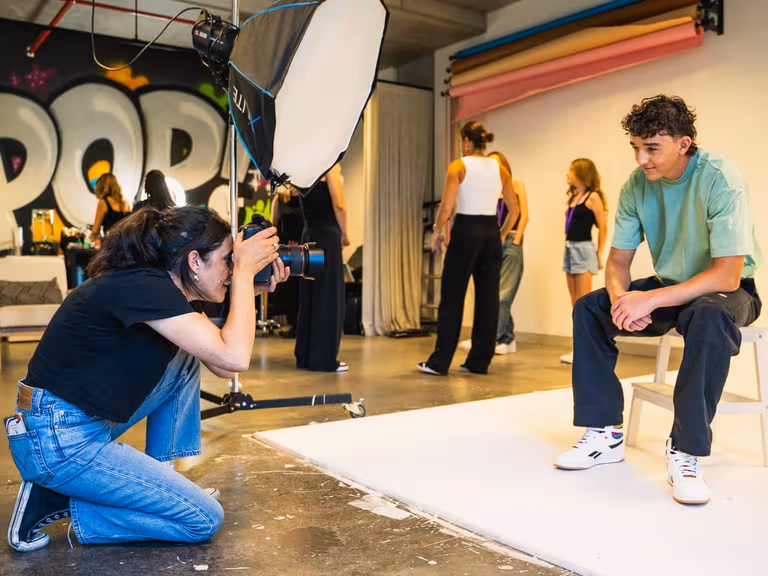 A POP Photography photographer crouches with a camera to shoot a teenage boy seated on a stool against a white studio backdrop, with a large softbox and a graffiti POP mural behind her