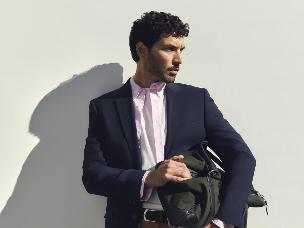 A man in a navy blazer, pink Sportscraft Oxford shirt and tan shorts holding a green weekender bag against a white wall