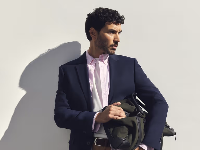 A man in a navy blazer, pink Sportscraft Oxford shirt and tan shorts holding a green weekender bag against a white wall