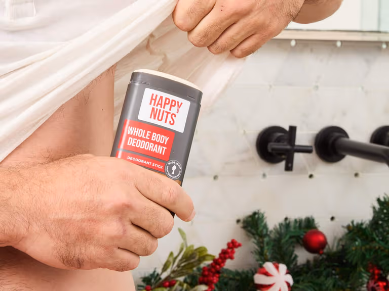 A man lifting his white shirt and holding a Happy Nuts Whole Body Deodorant stick against his side in a white tiled bathroom, with Christmas garland, red baubles and peppermint decorations on the bench behind him.