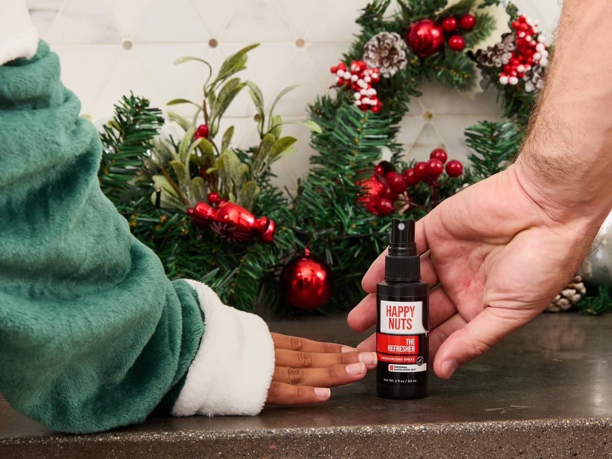 A hand setting down a black bottle of Happy Nuts The Refresher deodorising spray on a stone bathroom counter in front of a Christmas wreath with red baubles and pine cones, beside a hand in a green fleece robe sleeve.