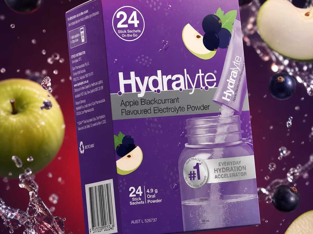 A purple 24 pack box of Hydralyte Apple Blackcurrant Stick Sachets surrounded by apple slices, blackcurrants and splashing water
