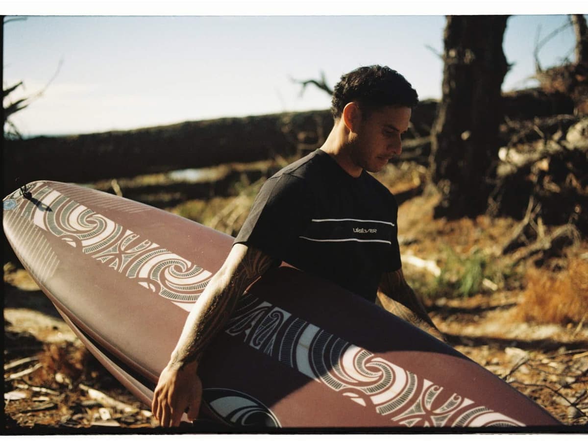 Kehu Butler carrying a SharpEye surfboard decorated with Tai Kehu artwork through coastal scrub