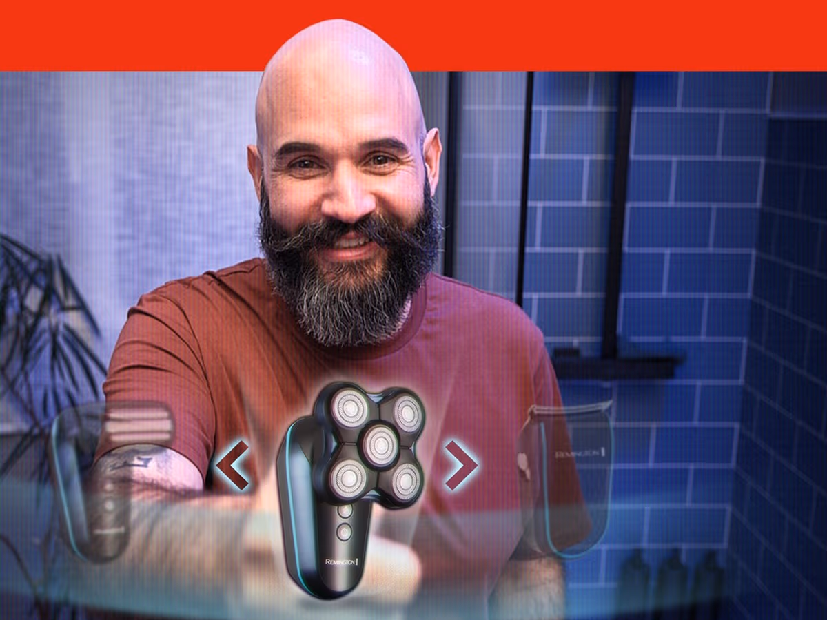 A bald man with a full dark beard holding the Remington Balder Head & Beard shaver towards the camera in a blue tiled bathroom