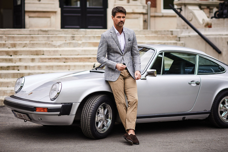 Man in a gray coat over white shirt and khakis standing with his back leaning on front fender of a Porsche 911